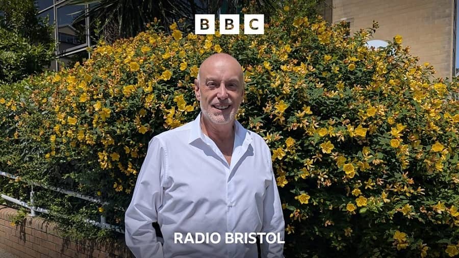 A smiling man in a white shirt stands before yellow flowers, with the BBC logo and "RADIO BRISTOL" text.